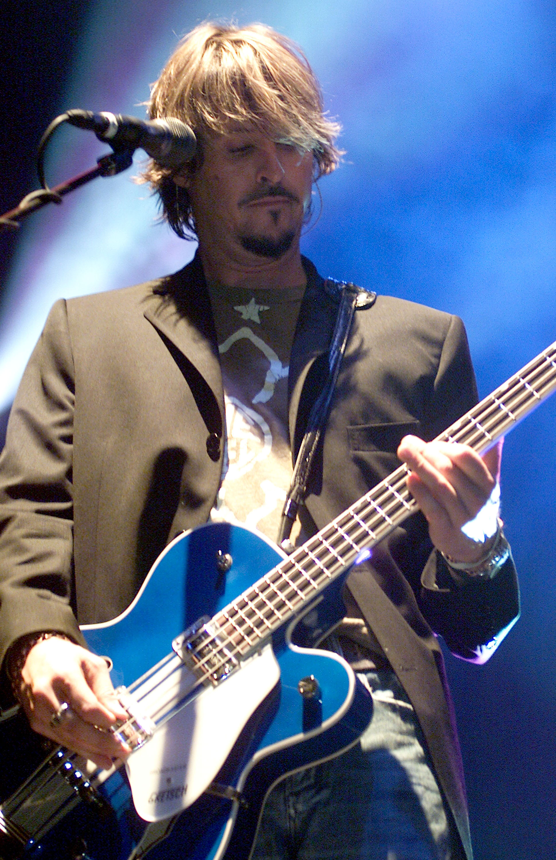 Charlie Colin from the rock band Train stands on stage wearing a suit jacket and playing a blue bass guitar