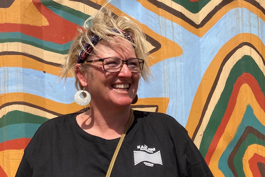 A smiling blonde woman in black t-shirt stands in front of a colourful painted backdrop. 