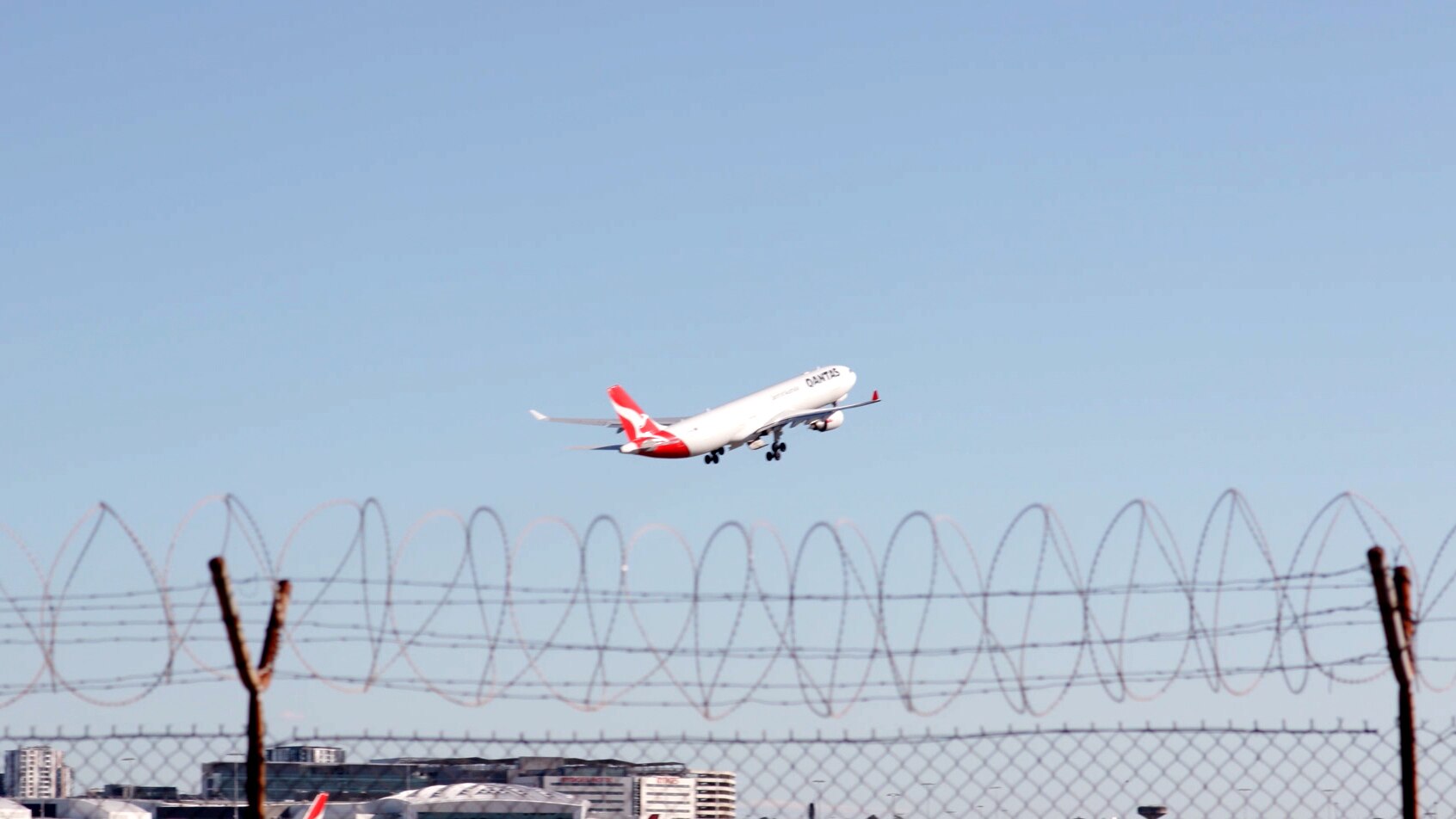 A Qantas plane takes off. In the foreground underneath the plane is the barbed wire of an airport fence.