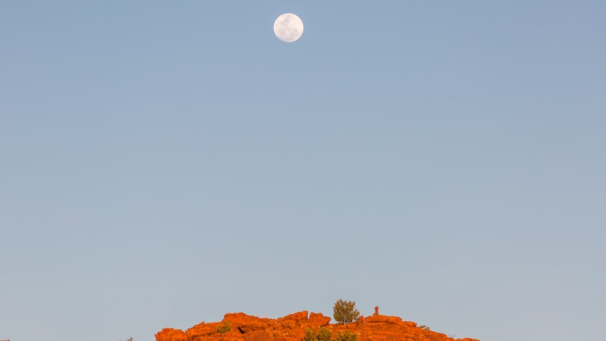 A full moon hangs in the sky above a red rocky formation.