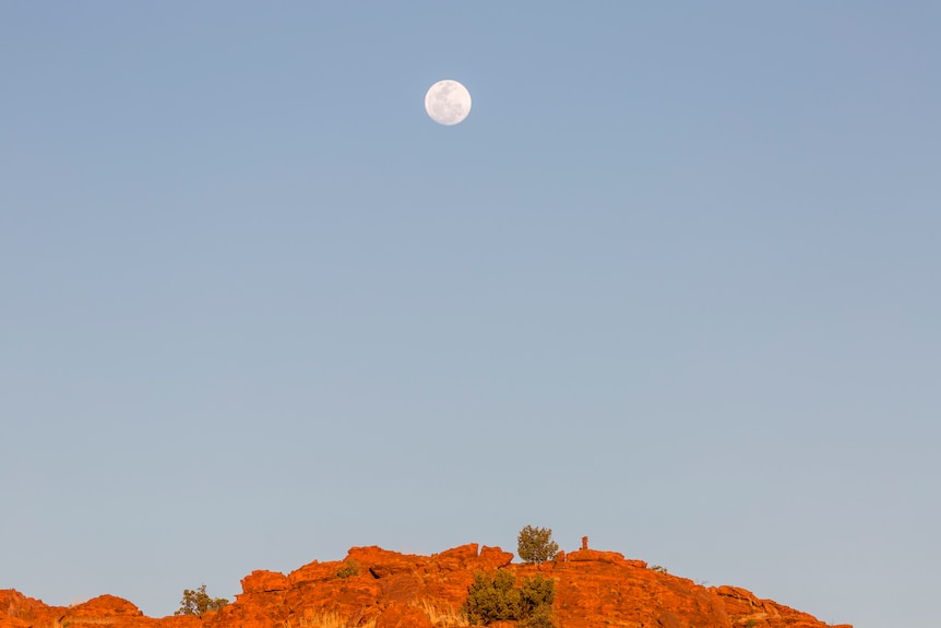 A full moon hangs in the sky above a red rocky formation.