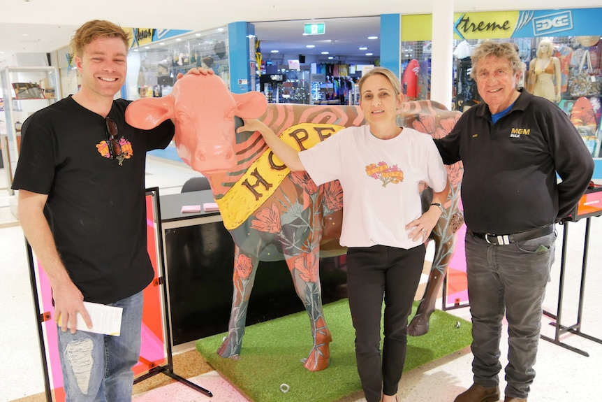 Three people stand in front of a cow sculpture which is painted pink and yellow with the word 'hope' visible. 