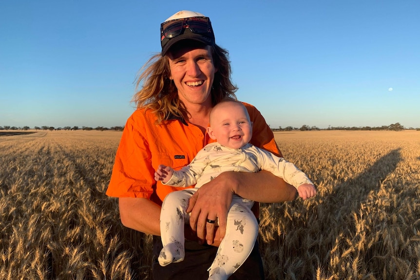A man wears an orange hi-vis shirt and a cap with sunglasses on it as he holds a smiling baby while standing in a field.