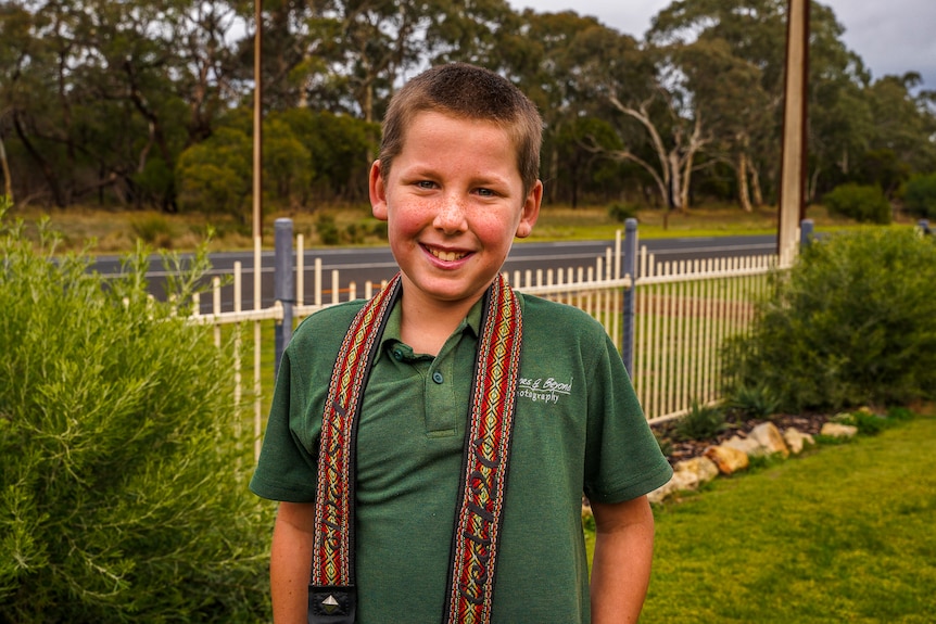 A smiling boy wearing a green polo shirt in a suburban front yard, his camera strap evident around his neck