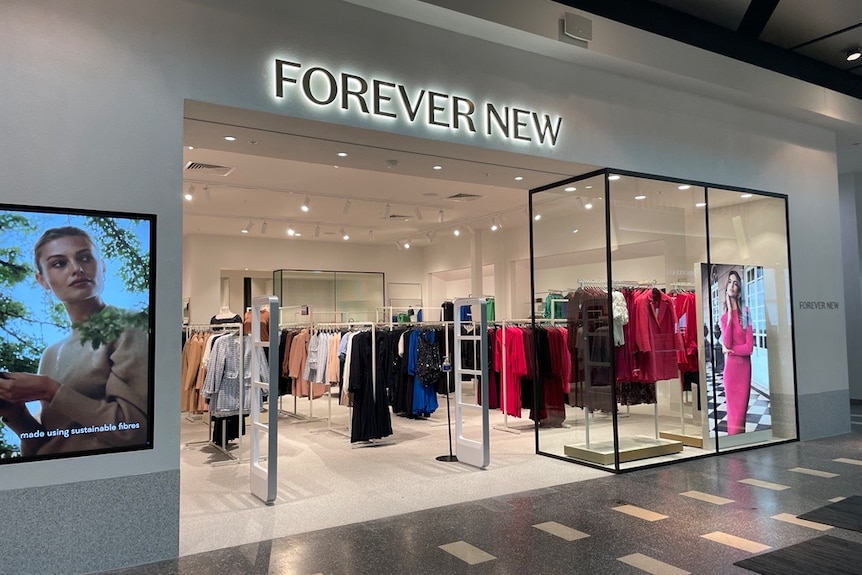 The front of a women's clothing store with the words Forever New lit up. Mannequins and clothing racks line the store.