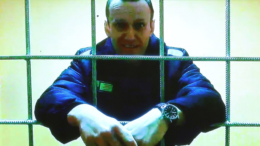 A close up of a man in a jail with his arms through the bars of the cell and then held together.