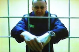 A close up of a man in a jail with his arms through the bars of the cell and then held together.