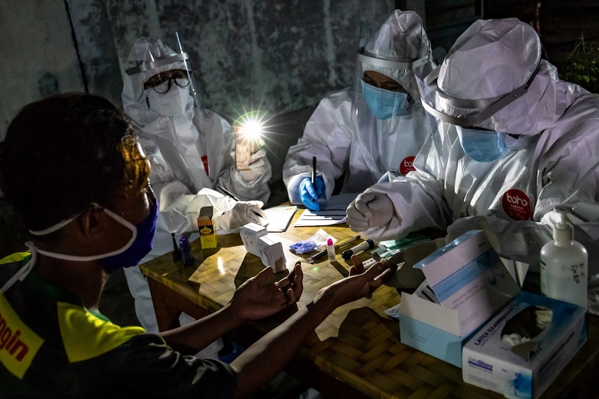 Three health workers in PPE conduct a test on a man under the light of an iphone torch