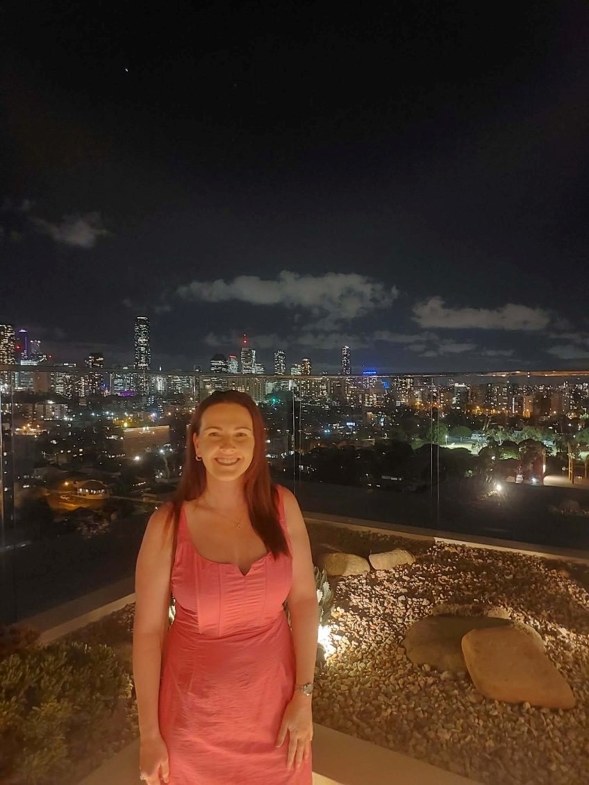 A woman in a pink dress standing on a balcony with city lights behind her. 