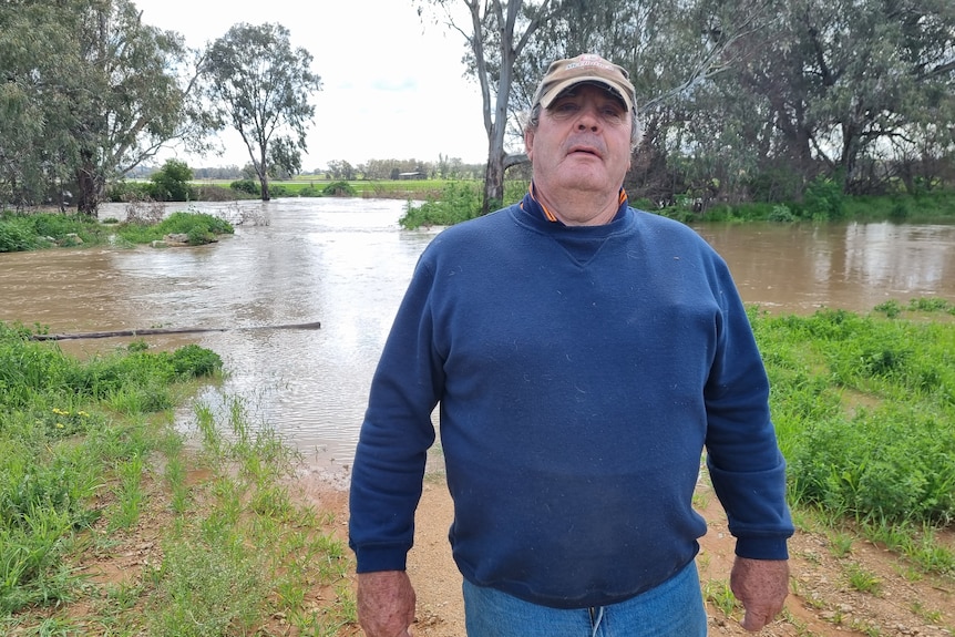 A man in a blue jumper and brown cap stands in front of a flooded river.