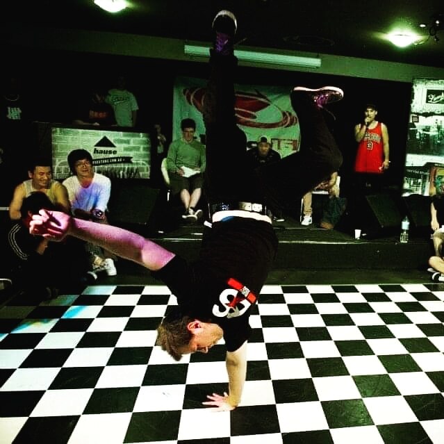 Man does a one-handed handstand on a black and white chequered dance floor.