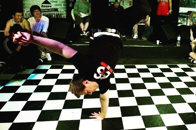 Man does a one-handed handstand on a black and white chequered dance floor.