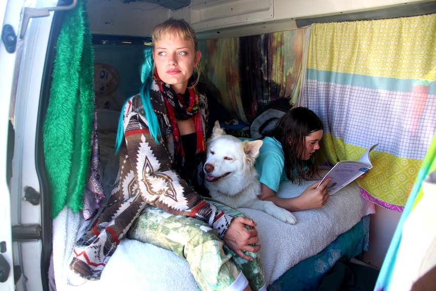 A mother with blonde hair sits next to her dog and daughter, who is reading a book