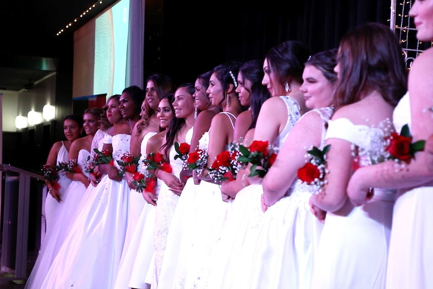 Indigenous debutantes line up in white gowns, holding sprays of red roses