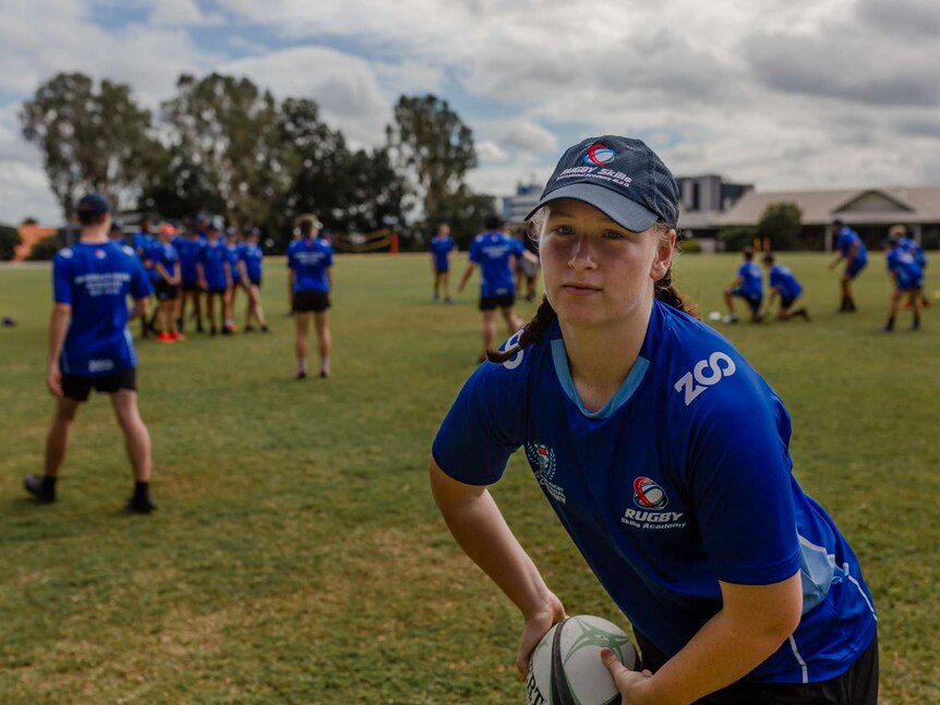 A young girl stares just off screen, about to toss a rugby ball under her arm. Oval and rugby players in background.