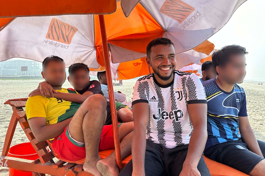 Samsul smiling, sitting under an orange and white umbrella with his friends. 