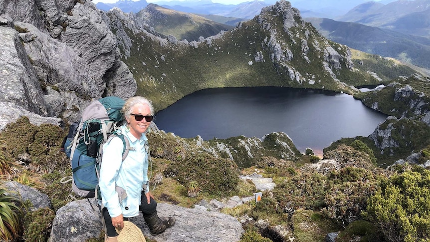 Mary, with white hair and sunglasses, smiles in front of a large lake surrounded by mountains.