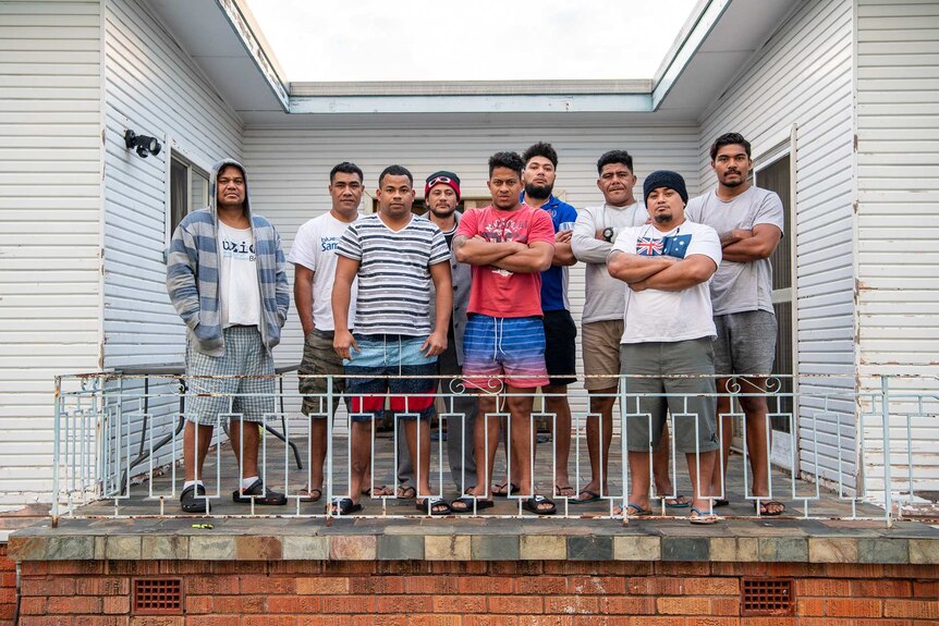 Nine Samoan men stand outside their home in Inverell, with serious expressions on their faces. Five cross their arms.