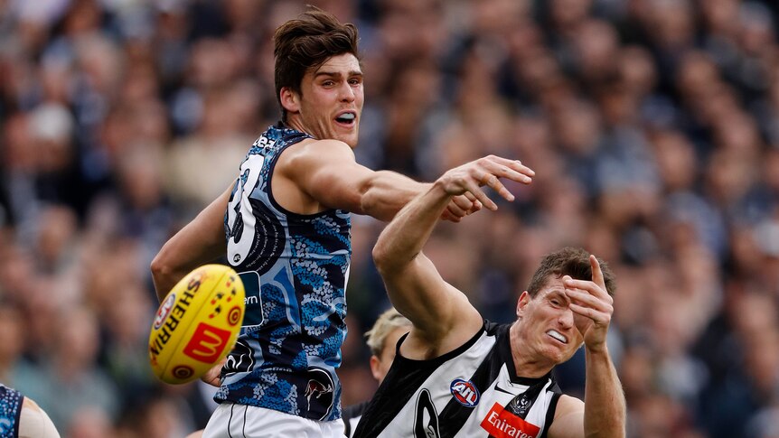 A Carlton defender extends his fist from an attempted spoil and a Collingwood forward grimaces as the ball flies away.