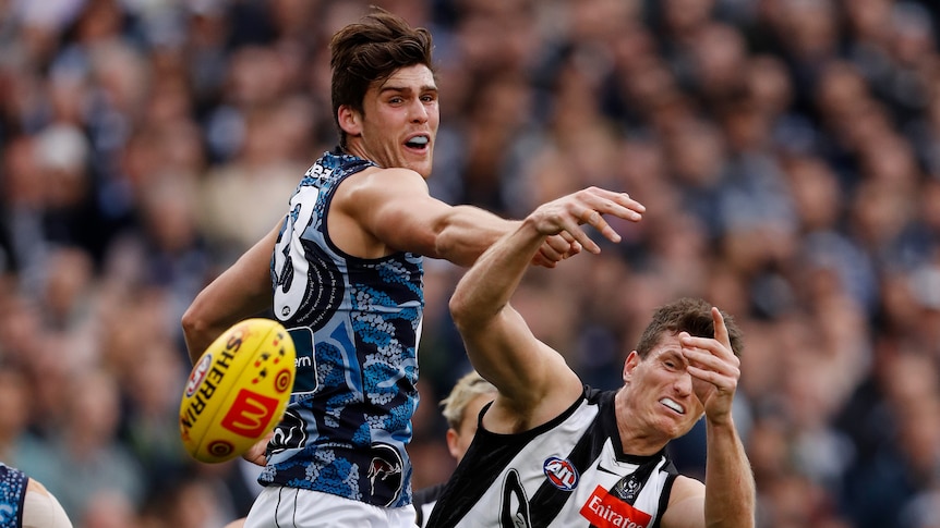 A Carlton defender extends his fist from an attempted spoil and a Collingwood forward grimaces as the ball flies away.