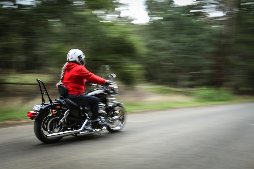 A woman in a red hoodie guns her motorcycle down the road, the forested background a-blur.