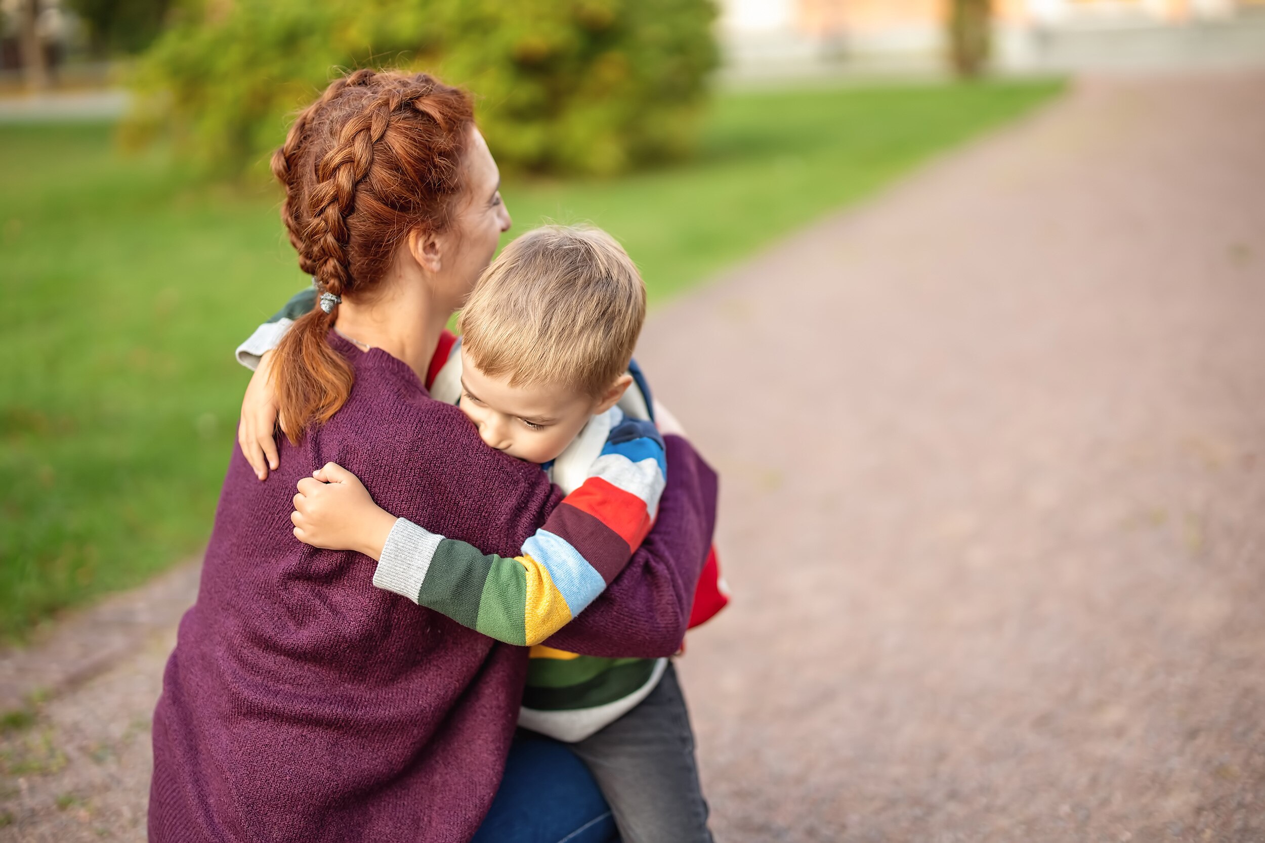 A woman with braided red hair kneels and embraces a young boy with blonde hair who looks upset.