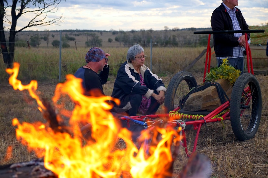 Two people sit, and another stands, by a campfire and a rock on a cart.