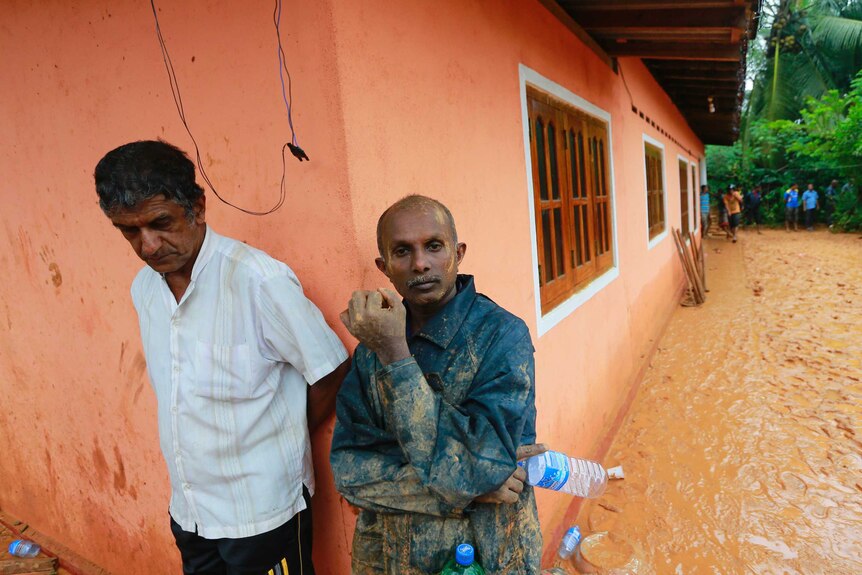 Two men lean against a house affected by a mudslide.