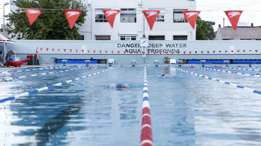 Various swimmers swim in a large pool, with a sign declaring 'danger deep water' on the back wall.