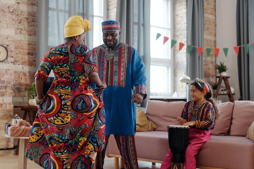Two adults in bright, colourful clothing dance together while a young girls plays a djembe drum.