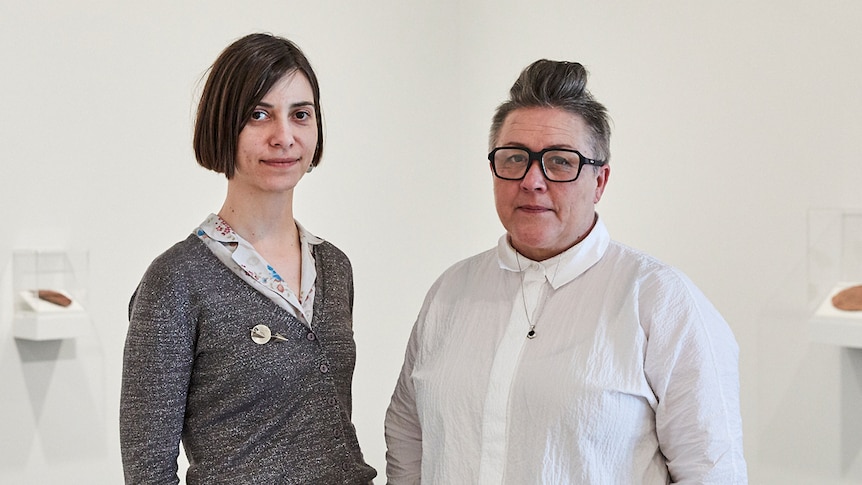 Young woman and middle-aged woman stand in white gallery space with objects in vitrines on wall behind.