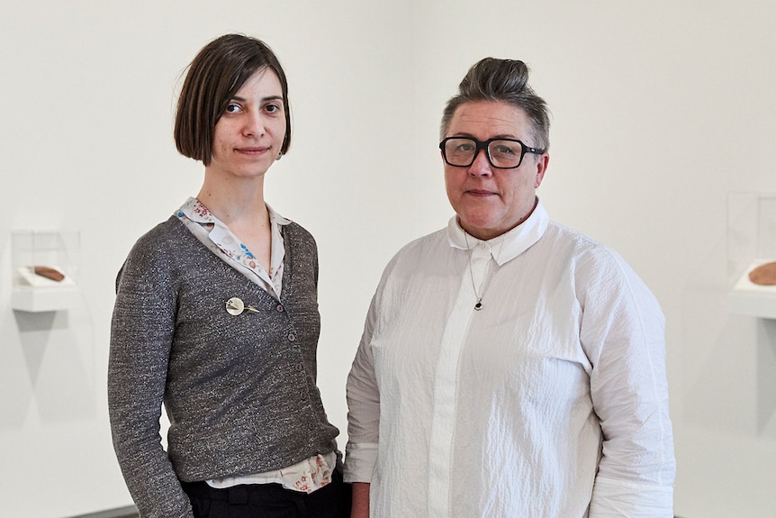 Young woman and middle-aged woman stand in white gallery space with objects in vitrines on wall behind.