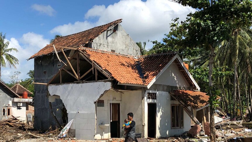 A man wearing a black shirt holds a baby as he walks past a wrecked house.