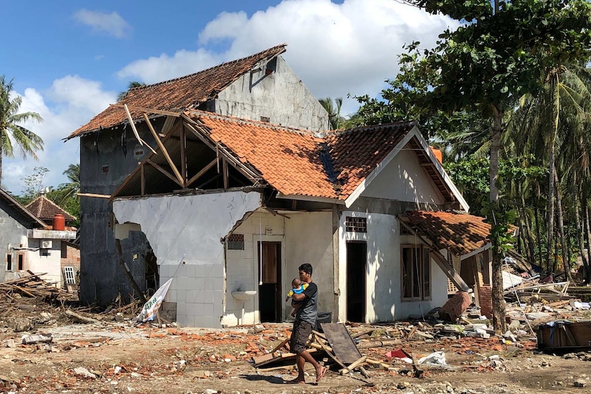 A man wearing a black shirt holds a baby as he walks past a wrecked house.