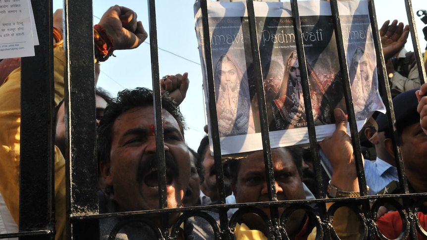 man yelling with one fist in the air and other holding sign with Padmaavat poster against gate, other protesters in background