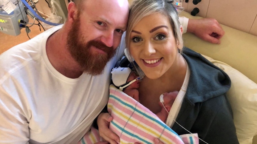 A bald man with a beard and a blonde woman holding two premature babies in a blanket