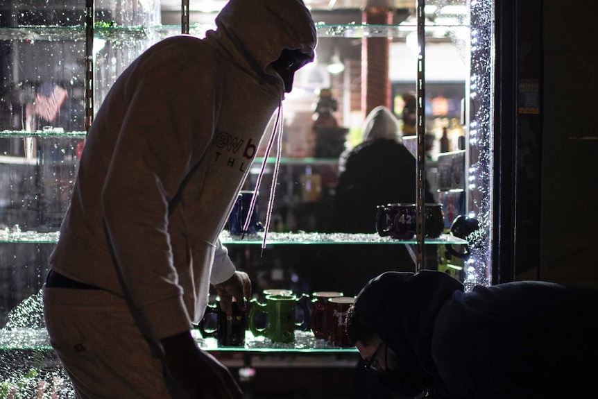 Three men in hoodies stand in front of broken glass in a dark store taking goods