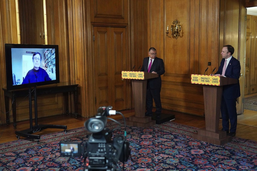 Two suited men stand behind lecterns 1.5 metres apart. A woman on a screen, joining the press conference by video, is visible.