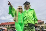 A woman and man in green jumpsuit and 1970s gear in front of a large country hotel