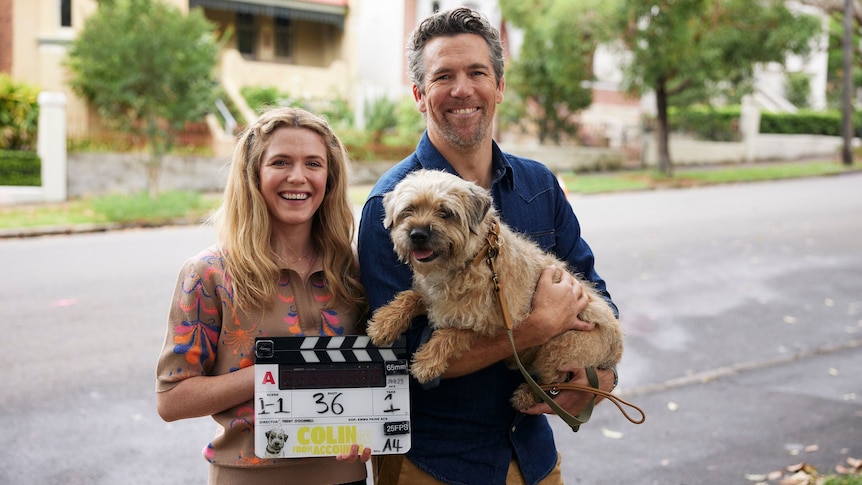 Harriet Dyer with a clapper board and Patrick Brammall holding a dog (Colin) standing outside on the street