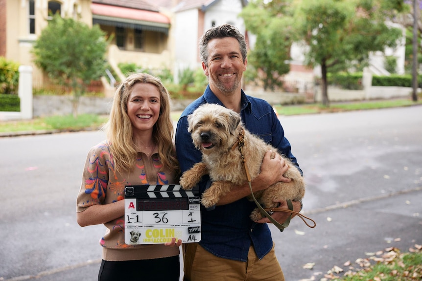 Harriet Dyer with a clapper board and Patrick Brammall holding a dog (Colin) standing outside on the street