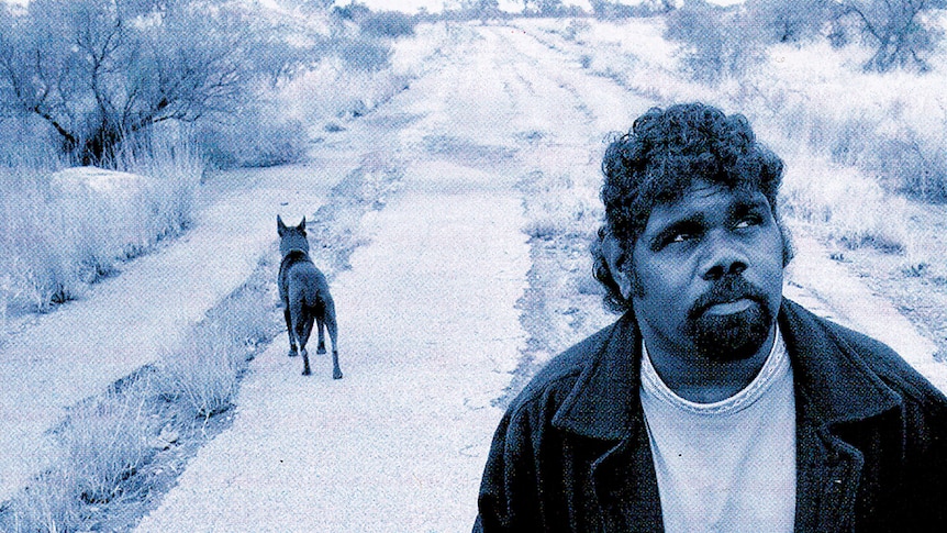 Frank Yamma looks into the distance on a deserted dirt road. A black dog behind him looks the other way.