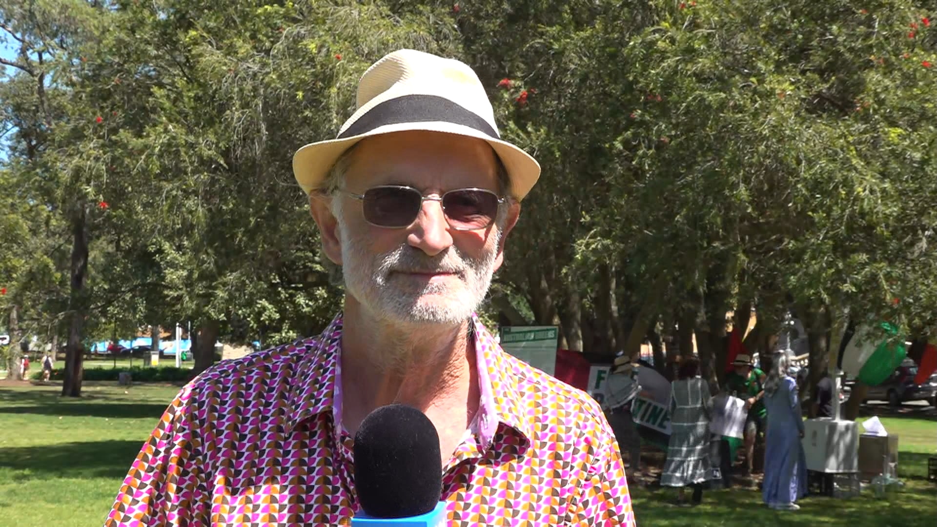 A man wearing a fedora and a pink patterned shirt speaks into a microphone at a park.
