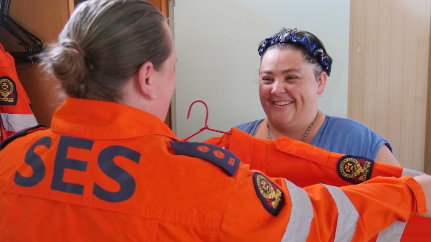 Woman holds orange state emergency service jacket up in front of other woman looking for size