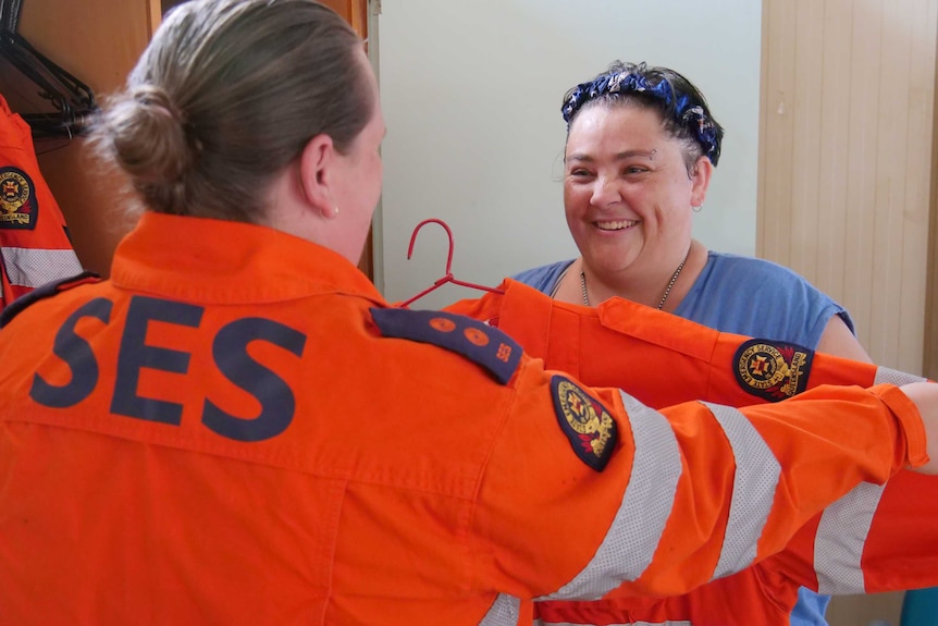 Woman holds orange state emergency service jacket up in front of other woman looking for size