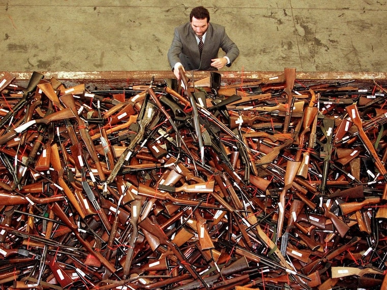 An aerial shot of a man in a brown suit stacking rifles on top of each other on a table.