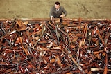An aerial shot of a man in a brown suit stacking rifles on top of each other on a table.