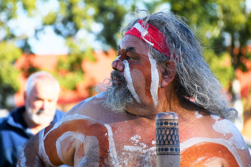 An Aboriginal man in traditional dress holds a didgeridoo and looks off to the side.