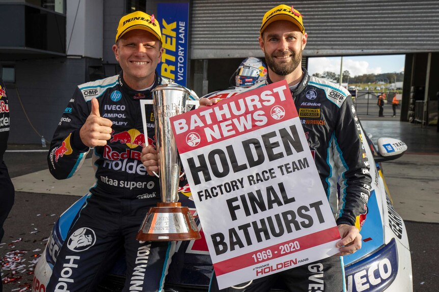 Двое улыбающихся мужчин в гоночной экипировке держат плакат с надписью о победе.