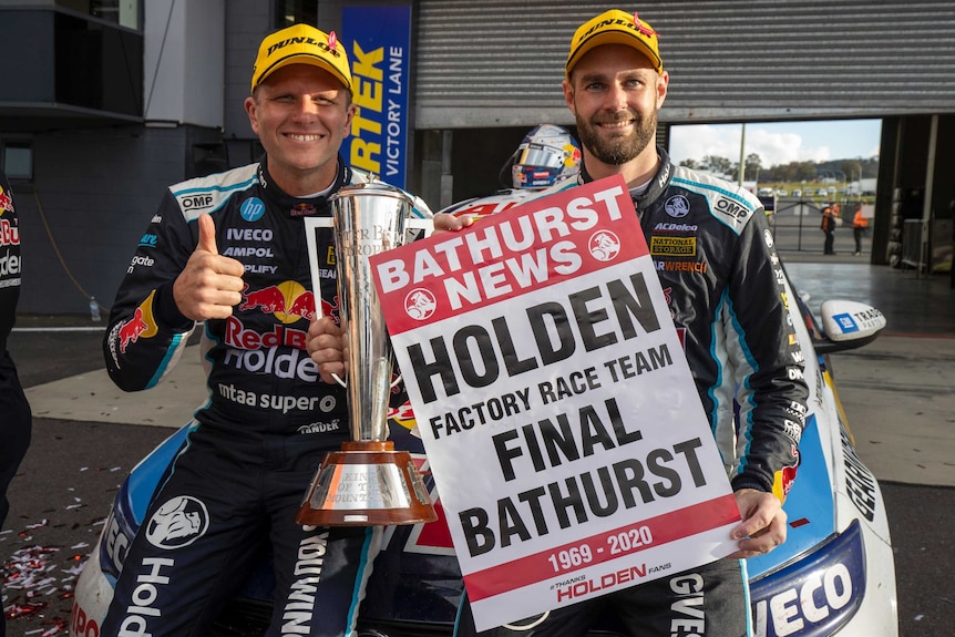 Two smiling men in motor racing gear hold up a poster marking a win.